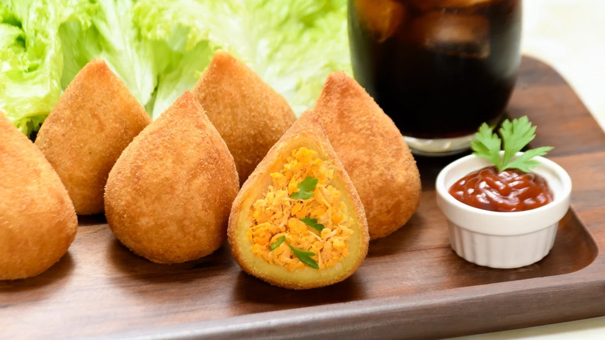 Imagem de porção de coxinhas de frango. Uma das coxinhas está aberta. Há um molho de ketchup ao lado da coxinha.